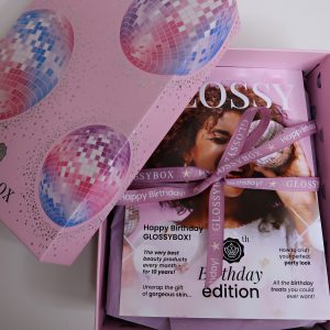10th Birthday Edition Glossybox: Pink cardboard box with the lid placed across one side revealing a magazine wrapped in a lilac ribbon