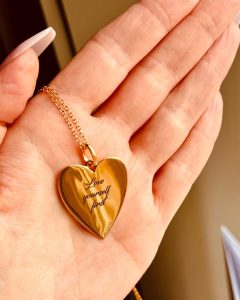 International Women's Day: A close-up of a gold heart-shaped pendant engraved with the words 'Love yourself first' held in a hand with a manicured finish.